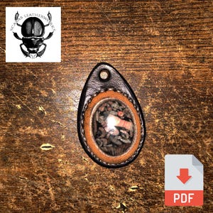 May include: A brown leather pendant with a round, clear window revealing a mix of small, colorful stones. The pendant is oval-shaped and has a black leather border.