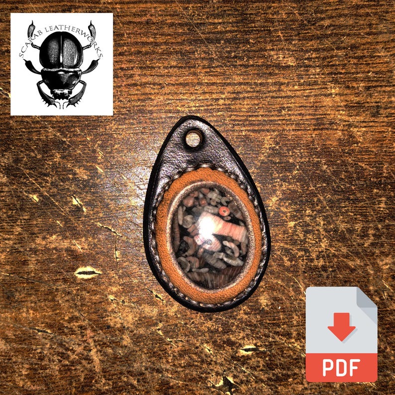 May include: A brown leather pendant with a round, clear window revealing a mix of small, colorful stones. The pendant is oval-shaped and has a black leather border.