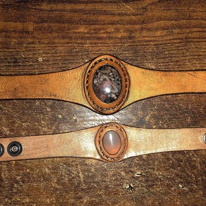 May include: Two leather bracelets with metal snaps and a round stone inlay. The top bracelet has a brown stone inlay and the bottom bracelet has a pink stone inlay.