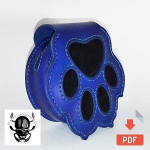 Puede incluir: Una bolsa de cuero azul en forma de huella de pata con forro de gamuza negro. La bolsa está cosida con hilo blanco. La bolsa es un proyecto artesanal y la imagen incluye un icono de descarga y la palabra "PDF".