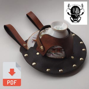 May include: A black leather tea cup holder with brown leather straps and accents. The holder has gold rivets around the edge and holds a white teacup and saucer with a floral design. The logo "SCARAB LEATHERWORKS" is in the upper right corner.