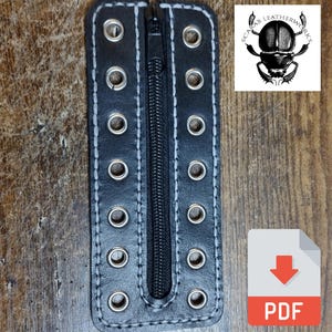 Puede incluir: Tira de cuero negro con una cremallera en el centro y ojales plateados en los bordes. La tira está cosida con hilo blanco. La tira está etiquetada con "Scarab Leatherworks".