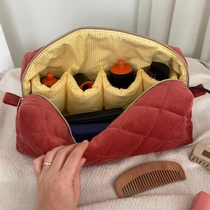 May include: A red corduroy cosmetic bag with a yellow gingham lining. The bag is open, revealing compartments holding hair styling tools. A wooden comb and hair clips are next to the bag. The bag has a zipper closure and a handle.