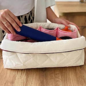 May include: A white quilted storage basket with a red and white gingham pattern. The basket contains a blue hair straightener and other beauty accessories. The basket is on a wooden surface.