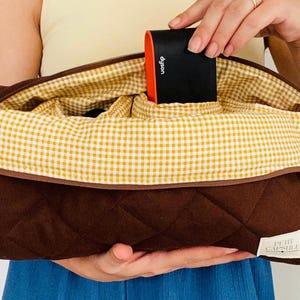 May include: A brown quilted cosmetic bag with a yellow and white gingham lining. The bag is open, revealing the interior, and a black and orange Dyson product is placed inside. The bag has a zipper closure and a small label that says "PETIT CAPSULE".