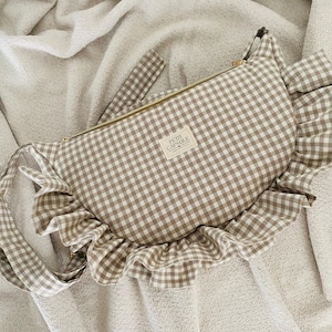 May include: A brown and white gingham fanny pack with a gold zipper and a ruffled edge. The bag has a matching strap and a small label that reads "PETIT CAPSULE". The bag is on a white surface.