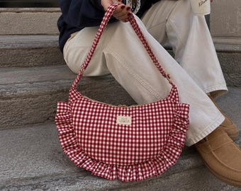 May include: A crescent-shaped shoulder bag featuring a red and white gingham pattern with ruffled fabric trim along the bottom edge. The bag includes a matching fabric strap and a small rectangular label on the front that reads PETITE CAPSULE.