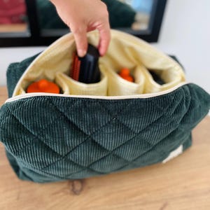 May include: Dark green quilted corduroy bag with a cream-colored interior and multiple compartments. The bag is open, revealing orange and black objects inside. A hand is reaching into the bag. The bag is sitting on a wooden surface.