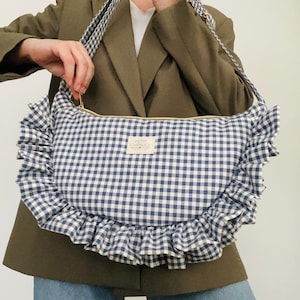 Op de afbeelding: Schoudertas met blauw-wit ruitpatroon met een gerimpelde rand. De tas heeft een gouden rits en een bijpassende ruitjesband. Het label op de tas luidt "PETITE CAPSULE".