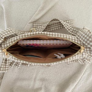 May include: A beige and white gingham tote bag with a gold zipper, open to reveal a pink floral notebook, pens, and other items. The bag has a ruffled edge and a matching gingham strap.