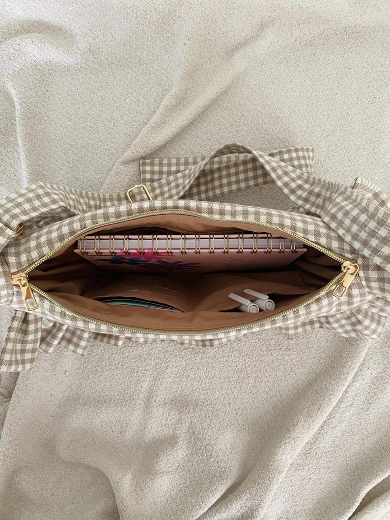 May include: A beige and white gingham tote bag with a gold zipper, open to reveal a pink floral notebook, pens, and other items. The bag has a ruffled edge and a matching gingham strap.