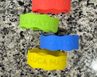 Könnte beinhalten: Vier farbige Silikon-Armbänder mit den Namen "Luca Mateo" und "Mateo" in Prägung. Die Armbänder sind in Rot, Grün, Blau und Gelb.
