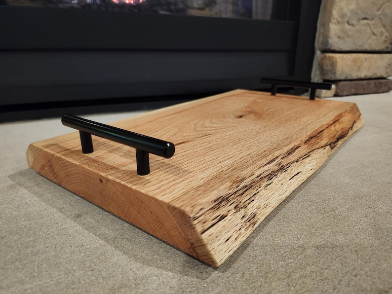 May include: A wooden serving tray with a natural wood finish and two black metal handles. The tray has a live edge design and is made from reclaimed wood.