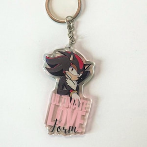 May include: A clear acrylic keychain featuring a cartoon character with black and red quills, wearing a black jacket and white gloves. The keychain has a pink rectangular base with the words "ULTIMATE LOVE Form" in white.