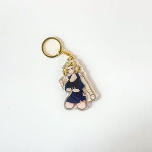 May include: A clear acrylic keychain featuring a blonde anime character in a fighting pose. The character wears a black top, a dark blue skirt, and a tan jacket. The keychain has a gold-colored ring.