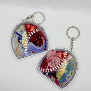 Op de afbeelding: Twee acryl sleutelhangers met zilveren sleutelringen. Elke sleutelhanger heeft een glas-in-lood ontwerp met een stripfiguur. Eén heeft een blauw-wit karakter, de andere een roze-rood karakter. Op een witte achtergrond.