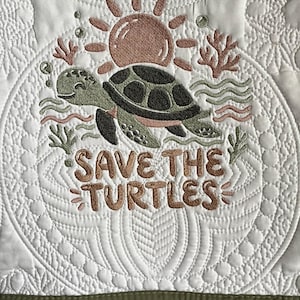 May include: White quilted fabric with an embroidered design featuring a smiling sea turtle, sun, and coral. The text "SAVE THE TURTLES" is embroidered in brown thread. The bottom edge is a solid olive green.