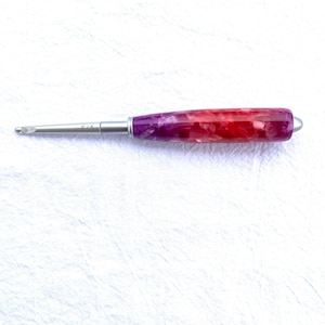 May include: A crochet hook with a stainless steel shaft and a colorful handle. The handle is a mix of red, pink, and purple hues. The hook has a size marking of F/5.