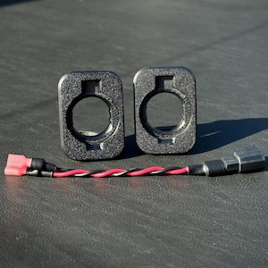 Puede incluir: Dos conectores eléctricos negros rectangulares con una abertura circular y una superficie texturizada. Un cable con hilos rojos, negros y blancos conecta los conectores, terminando en un conector rosa.