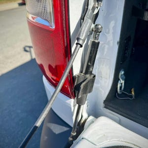 May include: Close-up of a white vehicle's rear tailgate with a black gas strut and a black cable attached to the tailgate.