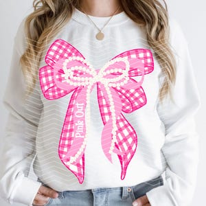 Op de afbeelding: Een wit sweatshirt met lange mouwen en een grote afbeelding van een roze met wit geruite strik, versierd met een snoer van witte parels. De tekst Pink Out is in het wit gedrukt op een van de linten van de strik, gecentreerd op de borst.