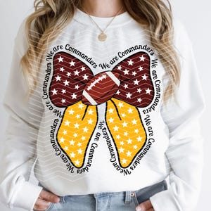 May include: White sweatshirt with a graphic of a football-shaped bow. The bow is burgundy with white stars, and yellow with white stars. The football in the center is brown and white. The words "We are Commanders" are written around the bow.