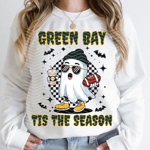 May include: White sweatshirt with a graphic of a ghost wearing sunglasses, a beanie, and yellow shoes, holding a football and a coffee. The text "GREEN BAY" and "TIS THE SEASON" are printed in green and yellow.