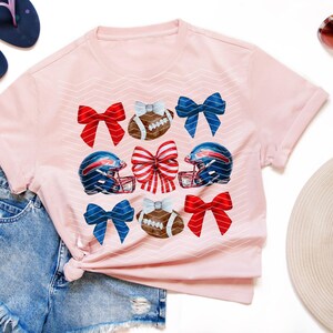 May include: Pink t-shirt with a design of football helmets, footballs, and red, white, and blue bows.