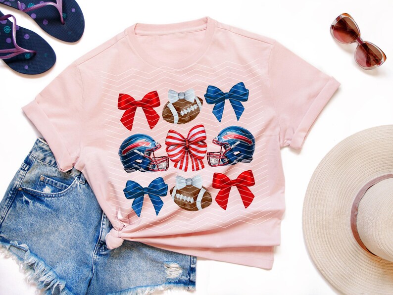 May include: Pink t-shirt with a design of football helmets, footballs, and red, white, and blue bows.