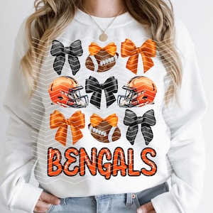 May include: White sweatshirt with the words "BENGALS" in orange letters. The sweatshirt has a pattern of orange and black bows and football helmets.