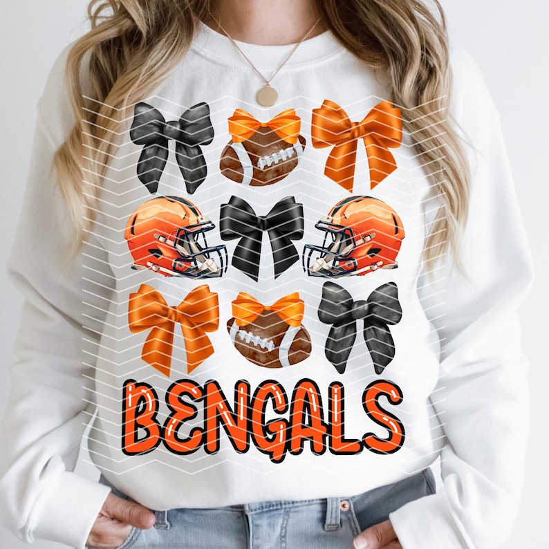 May include: White sweatshirt with the words "BENGALS" in orange letters. The sweatshirt has a pattern of orange and black bows and football helmets.