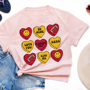 May include: A light pink t-shirt featuring heart-shaped candies with various designs. The candies have messages like "BE MINE", "LOVE YOU", and "XOXO". The shirt is paired with denim shorts, sandals, and sunglasses.