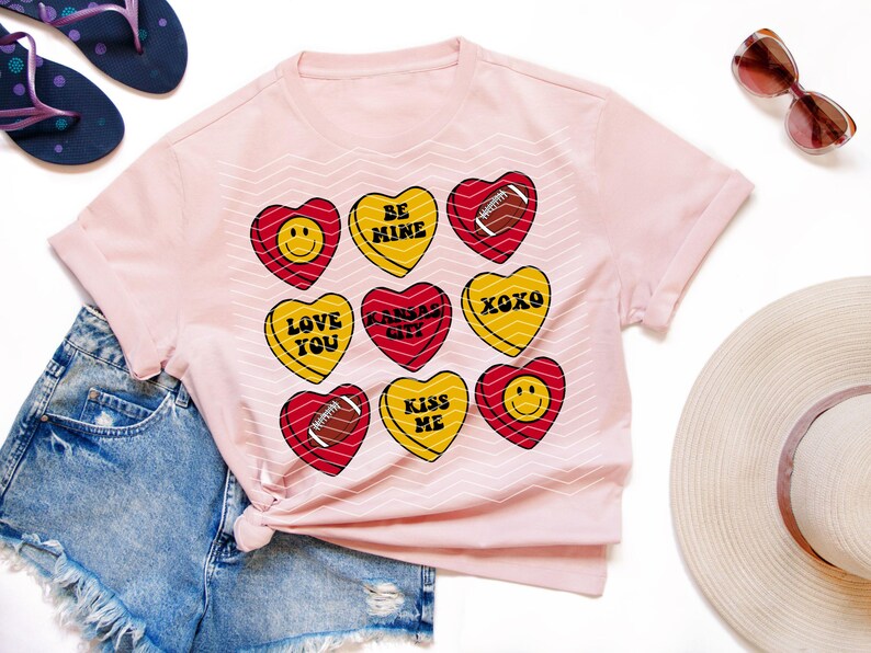 May include: A light pink t-shirt featuring heart-shaped candies with various designs. The candies have messages like "BE MINE", "LOVE YOU", and "XOXO". The shirt is paired with denim shorts, sandals, and sunglasses.
