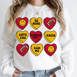 May include: White sweatshirt featuring a grid of nine heart-shaped designs. The hearts are red and yellow, with various graphics and text, including a football, smiley faces, and phrases like "Be Mine" and "Love You".