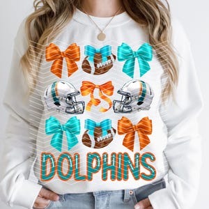 May include: White sweatshirt with a teal and orange design featuring football helmets, footballs, and bows. The text "DOLPHINS" is written in teal and orange.
