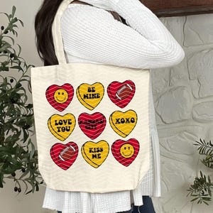 May include: Cream-colored tote bag featuring nine heart-shaped designs in red and yellow. Each heart has a different message, including "BE MINE," "LOVE YOU," and "KISS ME." Some hearts feature a football graphic or a smiley face.