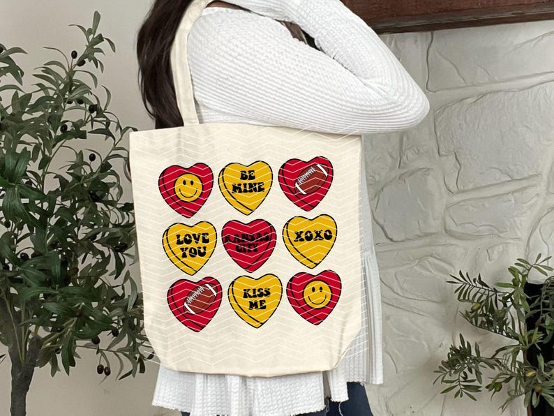 May include: Cream-colored tote bag featuring nine heart-shaped designs in red and yellow. Each heart has a different message, including "BE MINE," "LOVE YOU," and "KISS ME." Some hearts feature a football graphic or a smiley face.