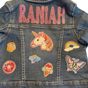 Peut inclure: Veste en jean bleue avec le nom "RANIAH" en lettres pailletées roses dans le dos. La veste est ornée d'écussons colorés, dont une licorne, un arc-en-ciel, un cupcake, un donut, un papillon, une abeille et une glace.