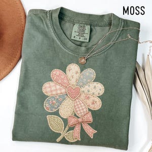 May include: A moss green t-shirt featuring a floral design with a patchwork pattern in shades of pink, blue, and cream. The flower has a heart-shaped center and a bow at the stem. A gold necklace with a small pendant rests on the shirt.
