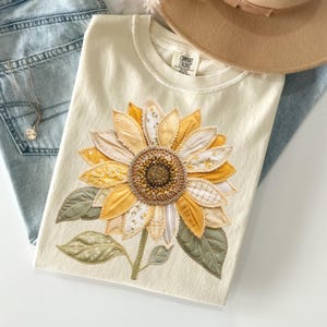 May include: Cream-colored t-shirt featuring a large, appliqued sunflower design. The sunflower has yellow, cream, and patterned fabric petals, a brown center, and green leaves. The shirt is folded, with a hat and jeans visible.