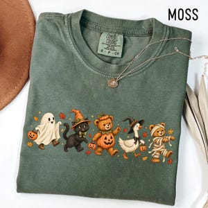 May include: A moss green crew neck t-shirt featuring a graphic of a ghost, a black cat in a witch hat, a teddy bear in a pumpkin costume, a goose in a witch hat, and a mummy teddy bear, all holding small pumpkins amidst scattered autumn leaves.