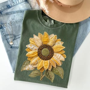 May include: Olive green t-shirt featuring a large, appliqued sunflower with yellow, cream, and brown fabric petals and green leaves. The shirt is paired with blue jeans and a tan hat. The shirt has a tag that reads XL 70-80.