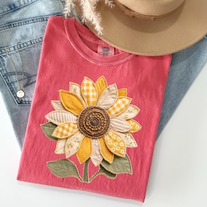 May include: A coral-colored t-shirt featuring a large, appliqued sunflower. The sunflower has yellow and cream-colored petals with gingham and floral patterns. A tan hat and blue jeans are partially visible in the background. A gold necklace is also visible.