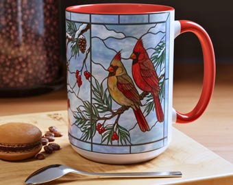 May include: White ceramic mug with a red interior and handle. The mug features a stained-glass style design of two cardinals perched on a branch with berries and pine needles. The background is a light blue with a snowy effect.