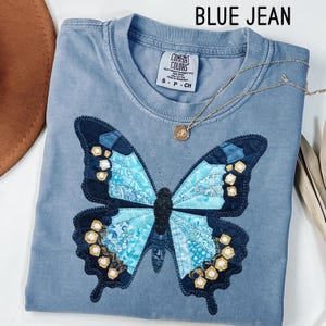 May include: A blue Comfort Colors t-shirt with a large butterfly applique. The butterfly features shades of blue and gold, with intricate patterns. A gold necklace with a pendant rests on the shirt. The text "BLUE JEAN" is at the top.