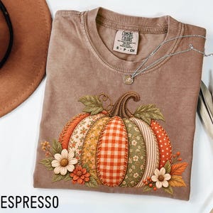 May include: An espresso-colored short-sleeve t-shirt featuring a large graphic of a pumpkin composed of various patterned fabric segments, including gingham, floral, and polka dot prints, accented with embroidered flowers and leaves.
