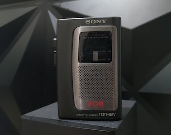 Puede incluir: Una grabadora de casetes Sony negra con una placa frontal plateada y letras rojas. El texto en la placa frontal dice "VO-R CASSETTE-CORDER TCM-82V".