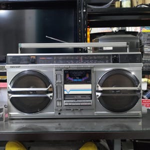 May include: Silver Sharp GF-800 boombox with large speakers, a radio dial, and a cassette player. The boombox has a retractable aerial and a VU meter. The speakers are black with silver accents. The boombox is on a glass surface.
