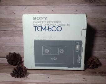 May include: A vintage Sony TCM-600 cassette recorder box. The box is light blue with the product name and details in black. A gray illustration of the recorder is on the front. The box is on a wooden surface.