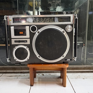 Może przedstawiać: Czarno-srebrny radiomagnetofon JVC z dużym centralnym głośnikiem, mniejszym głośnikiem poniżej, kieszenią na kasetę oraz wyświetlaczem tunera radiowego u góry. Urządzenie posiada srebrny panel sterowania oraz czarne uchwyty po bokach.